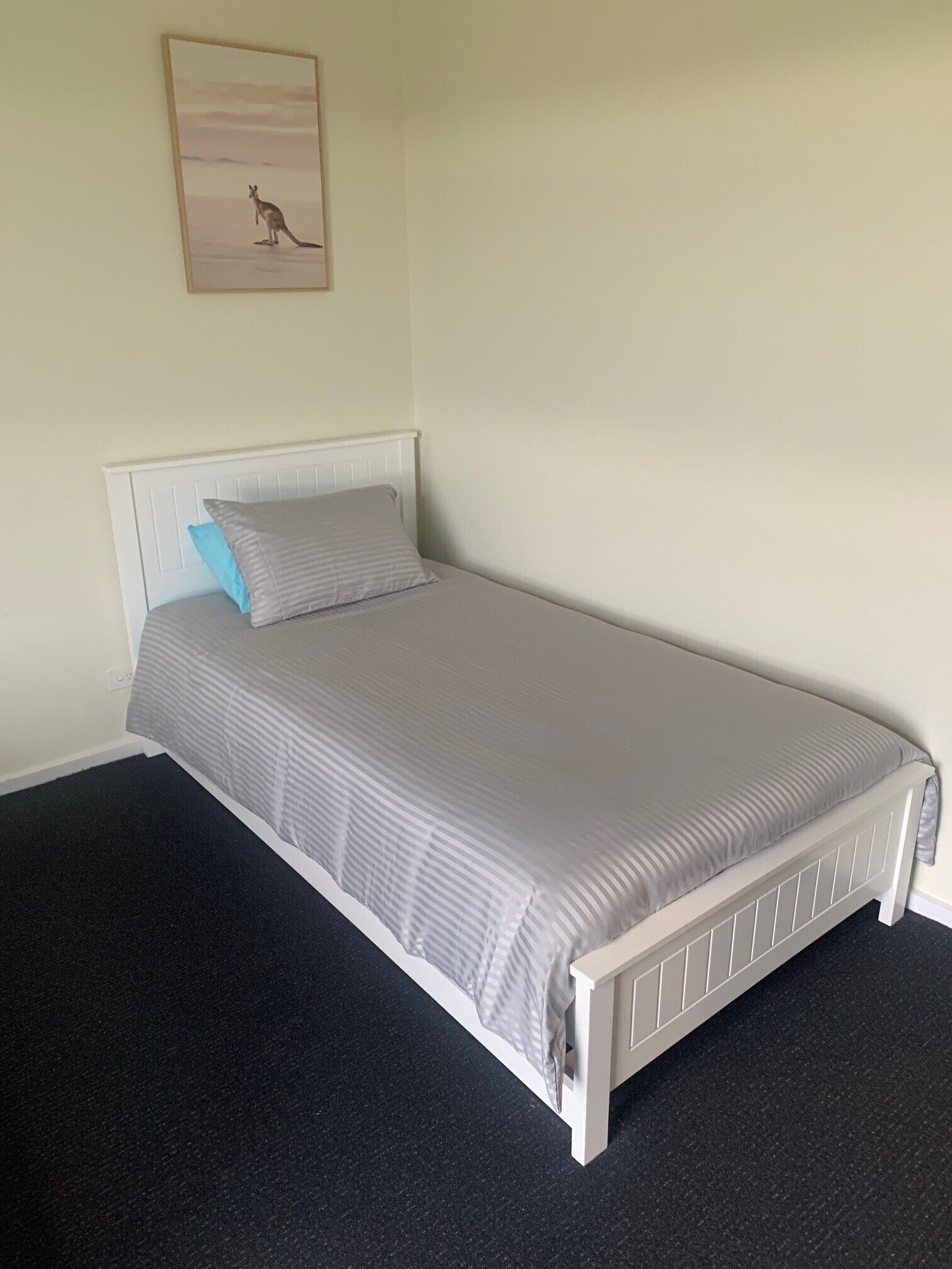 A single bed with a white headboard and grey bedding, a small blue pillow, and a framed picture of a kangaroo on the wall.