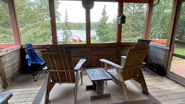 Een houten terras met twee Adirondack-stoelen en een kleine tafel, met uitzicht op een meer met bomen en een boot op de achtergrond.