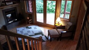 Een gezellig interieur van een chalet met een open haard, een houten trap, een geruit vloerkleed, een bekleedde bank en een raam met uitzicht op groen.
