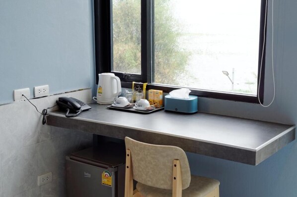 Un pequeño espacio de trabajo con un escritorio, una silla, una cafetera, un hervidor y una ventana con vistas a árboles.