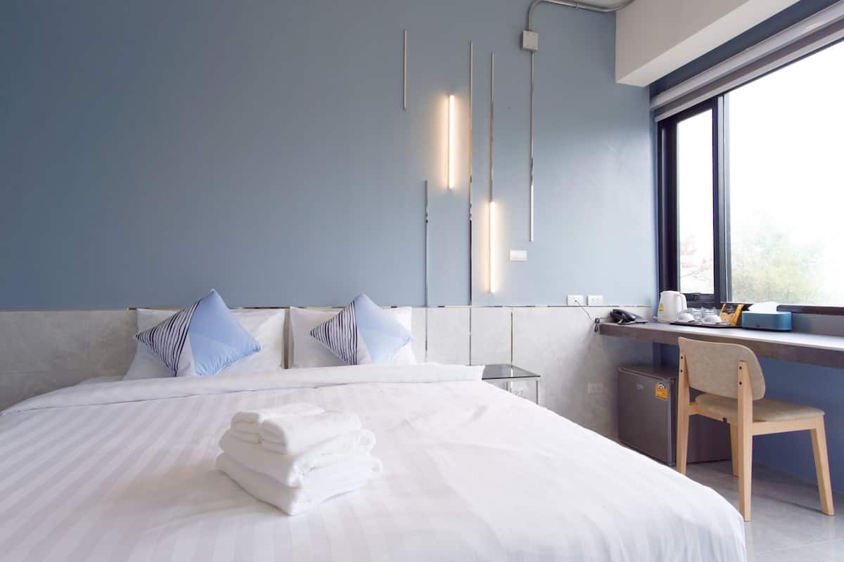 Une chambre d’hôtel moderne avec un grand lit, un bureau et une chaise. On y trouve deux appliques murales et une fenêtre donnant sur des arbres.