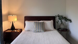 A neatly made bed with a checkered pillow, a bedside lamp, and a small table with a vase of green plants. - Newly Updated Home w/Easy Access to I-85/I-40  (Haw River)