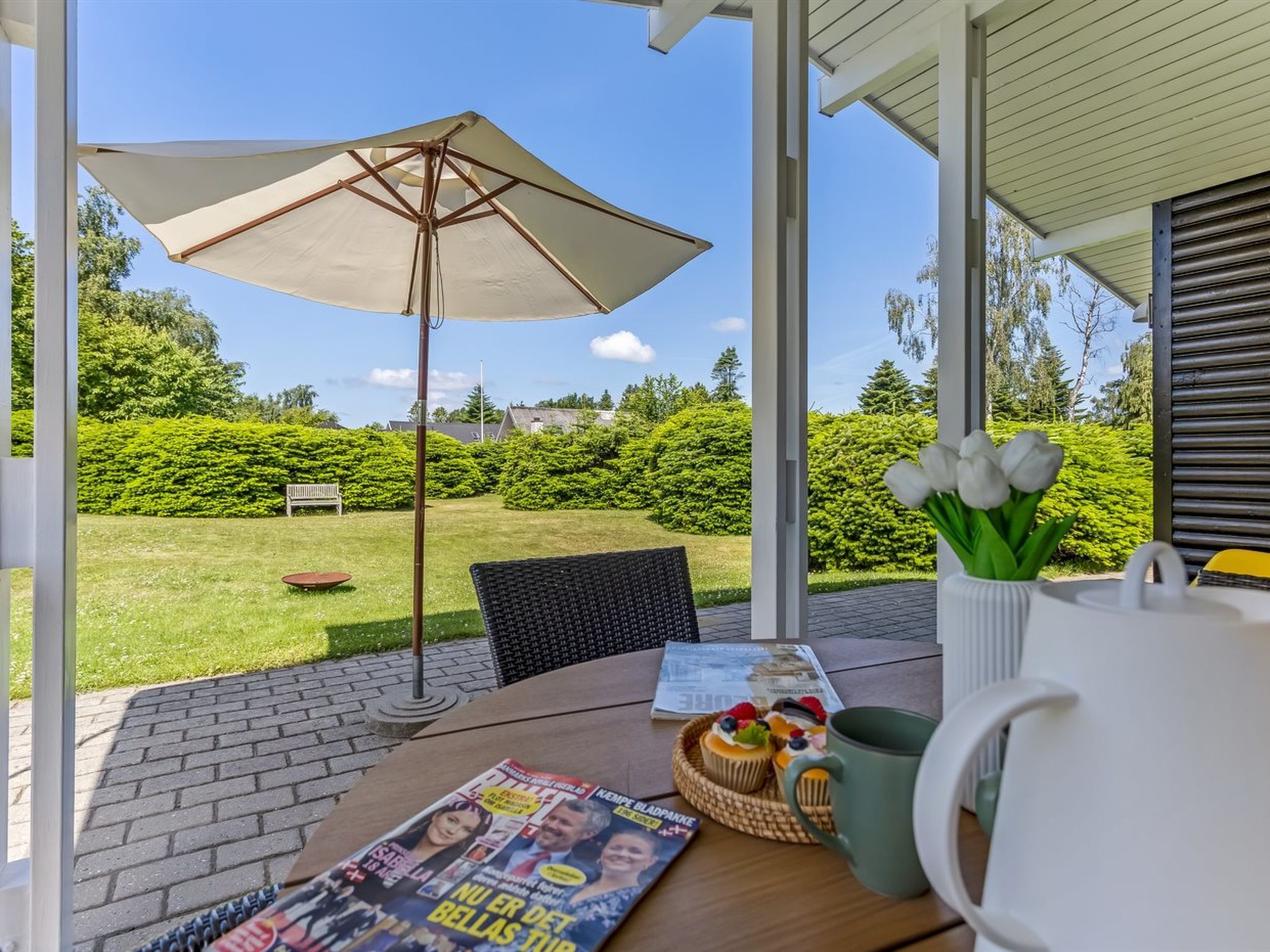 A patio with a table, a magazine, a basket of cupcakes, and a vase of flowers.