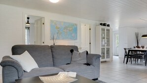 Uma sala moderna com um sofá cinzento, uma mesa de centro, uma área de jantar com mesa e cadeiras, e um armário branco com portas de vidro.