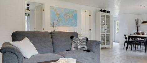 Una sala de estar moderna con un sofá gris, una mesa de centro, un comedor con mesa y sillas, y un mueble blanco con puertas de cristal.