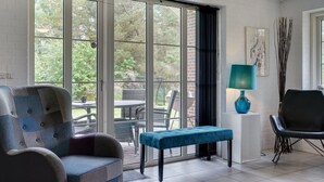 Quarto com uma grande porta de vidro deslizante que dá para um pátio externo, um banco estofado azul, uma cadeira preta e um abajur verde-água sobre uma mesinha de cabeceira branca.