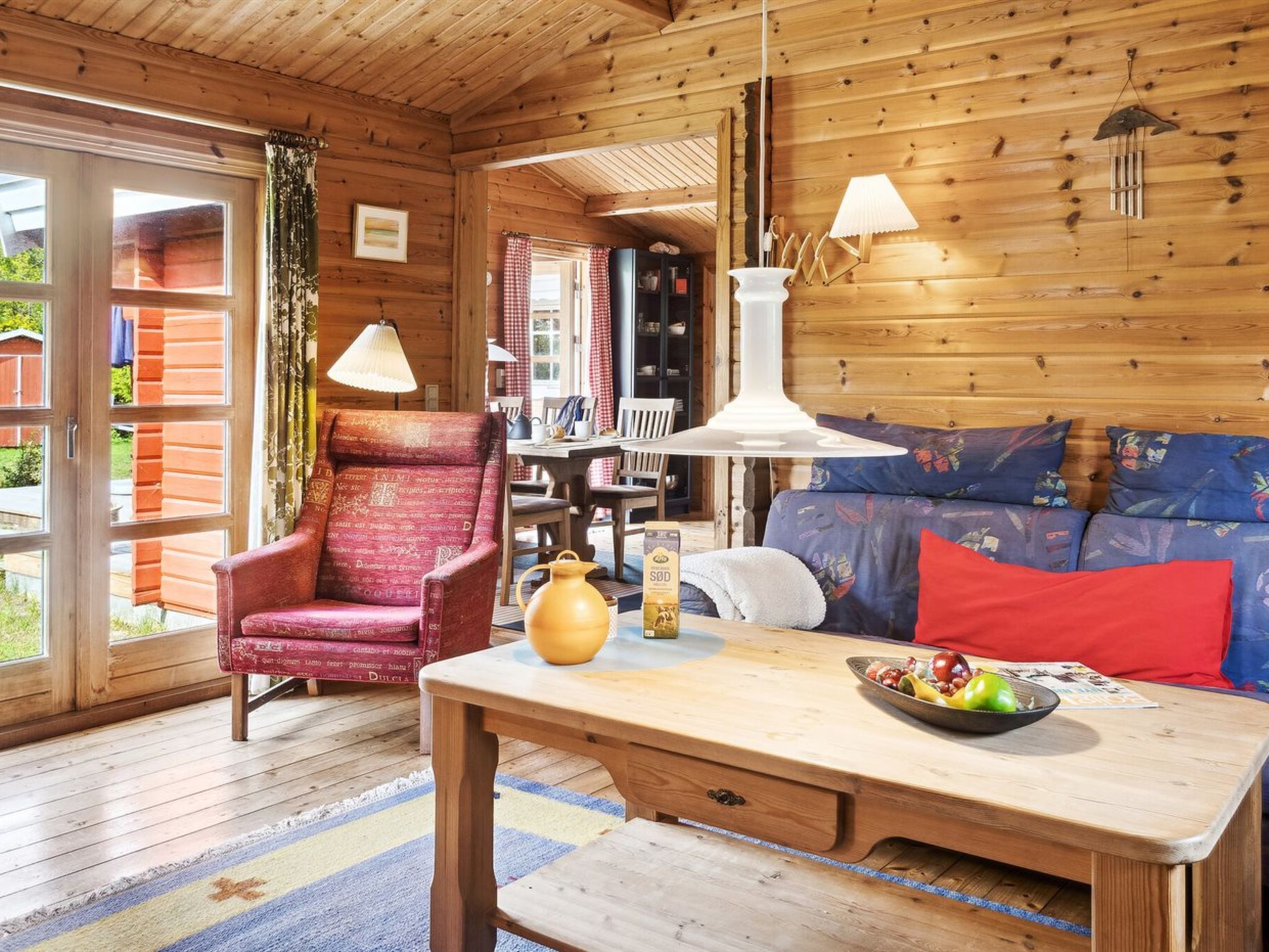 Ein gemütliches Blockhausschlafzimmer mit Holzbalkendecke, einem roten Sessel, einem Sofa mit bunten Kissen, einem Esstisch und einem großen Fenster mit Vorhängen.