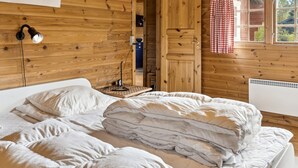 Een slaapkamer in een houten chalet met een bed, nachtkastje, lamp en een raam met geruite gordijnen.