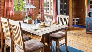 Een houten blokhut van binnen met een eettafel, stoelen, een lamp en een keukenhoek.
