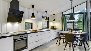 A modern kitchen with a built-in oven, a dining area with a round table and chairs, pendant lighting, and large windows with curtains. - "Aimi" - 270m from the sea by Interhome (Væggerløse)