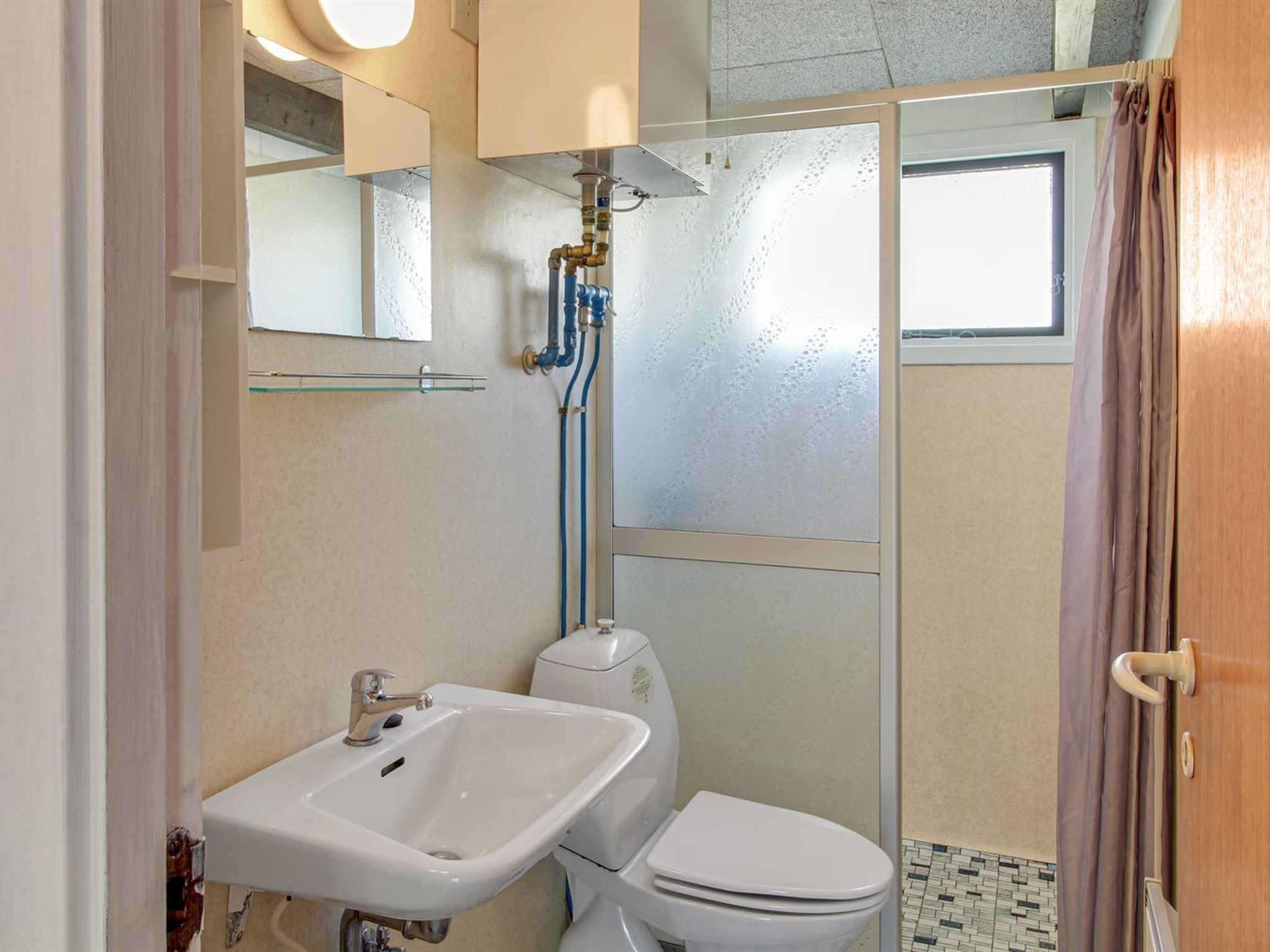 Bany amb un lavabo, vàter i dutxa de color blanc. Hi ha un mirall sobre el lavabo i una finestra sobre la dutxa.