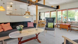 A modern living room with a sofa, a dining table, and a chair. There are hanging lights and a large window.