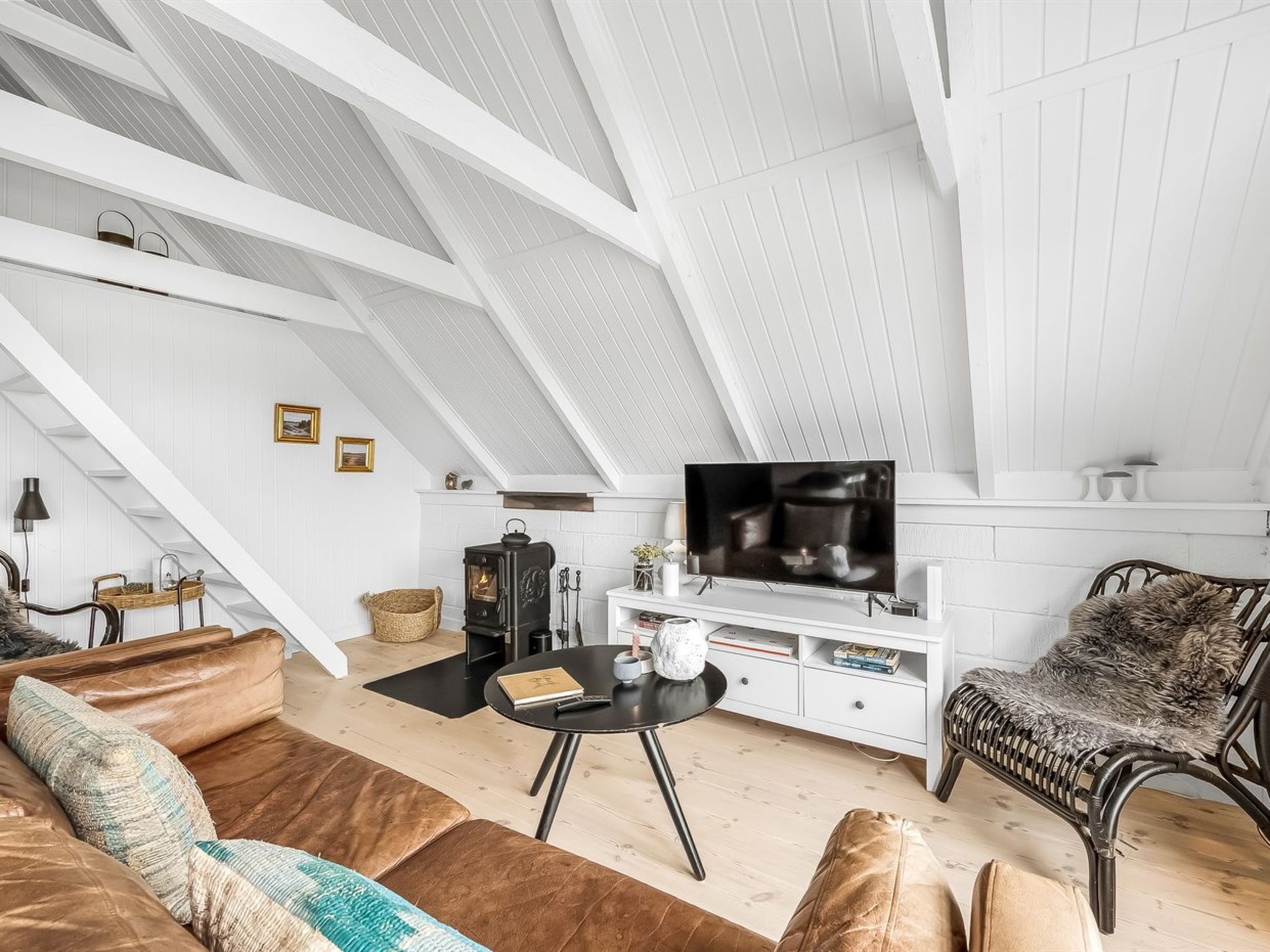 A loft-style living room with a high ceiling, white walls, a black TV on a white console, a brown leather sofa, a wicker chair, and a small table with a book and a candle.