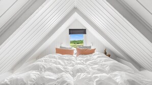Une chambre avec un plafond en pente, un lit avec une literie blanche, deux oreillers, une table de chevet avec une lampe et une fenêtre donnant sur un paysage verdoyant.