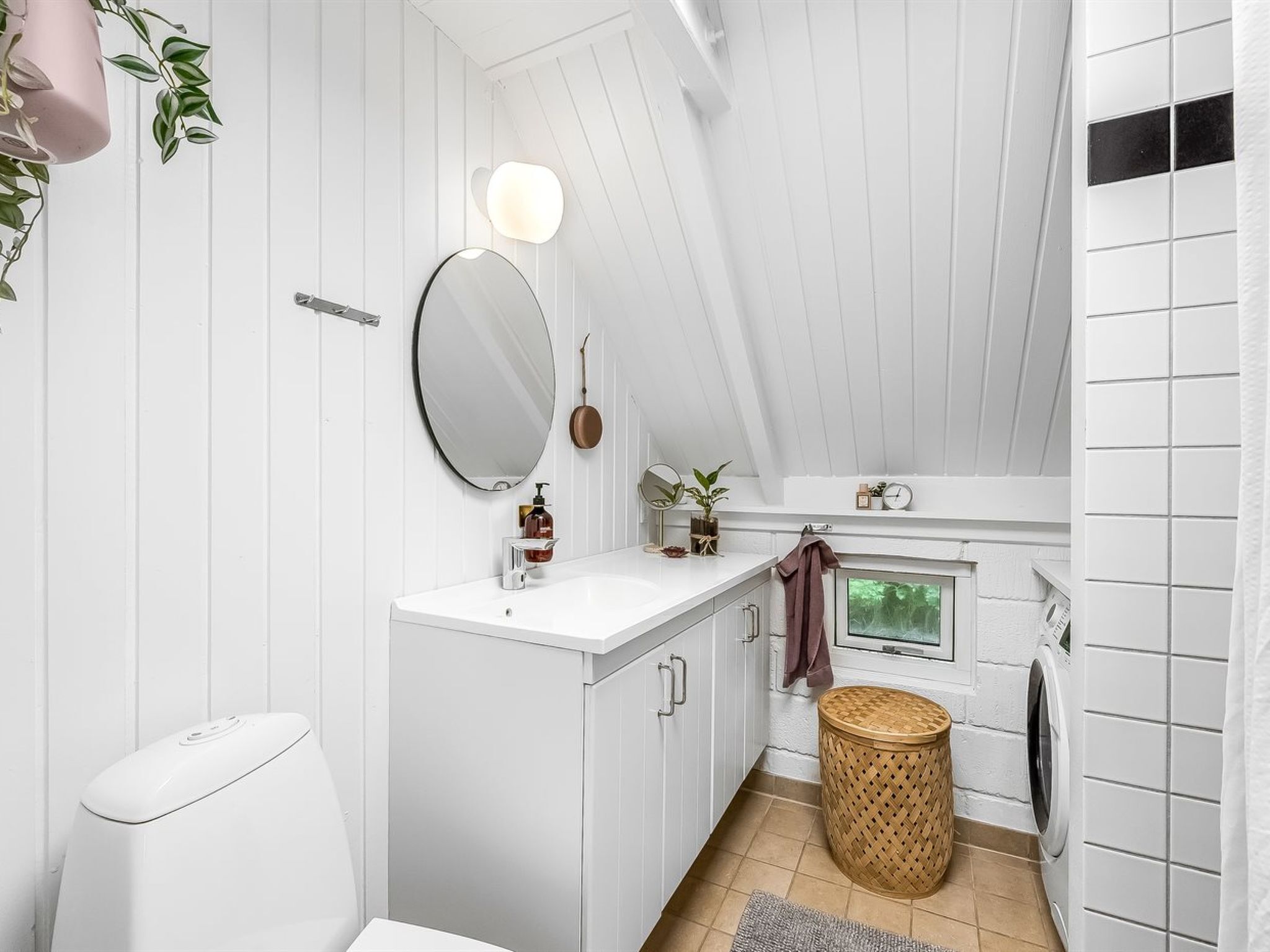 A modern bathroom with a white vanity, a round mirror, a small window, and a washing machine.