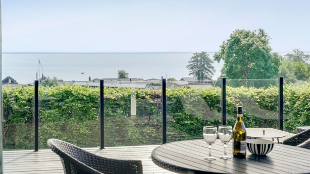 Une terrasse avec une table, des chaises et une bouteille de vin, donnant sur une étendue d’eau et un paysage verdoyant.