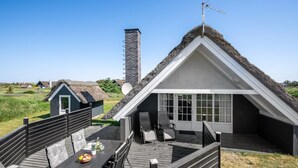 Une maison à toit de chaume dotée d’une extension moderne, une terrasse en bois avec mobilier d’extérieur et une vue dégagée sur le paysage environnant.