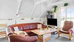 Un salon spacieux avec un canapé en cuir marron, une table basse en bois, un téléviseur et une grande fenêtre donnant sur l’extérieur.
