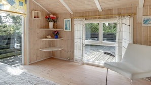 A wooden cabin interior with a white chair, wooden shelves with flowers, and a large window with curtains. - "Alfi" - 150m from the sea by Interhome (Sæby)