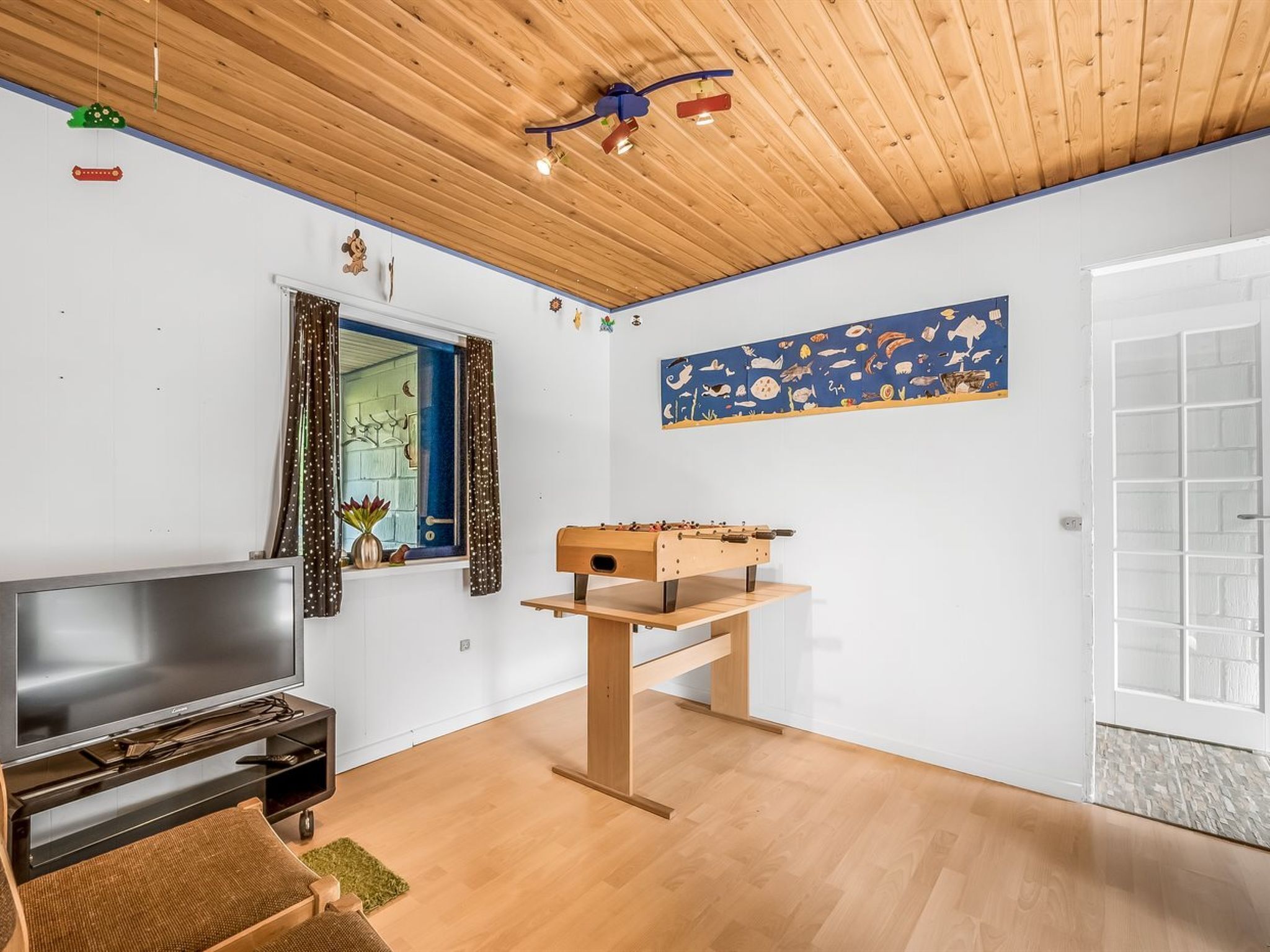 A modern living room with a wooden ceiling, a flat-screen TV, a foosball table, and a window with curtains.