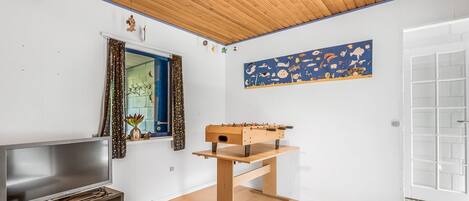 A modern living room with a wooden ceiling, a flat-screen TV, a foosball table, and a window with curtains.