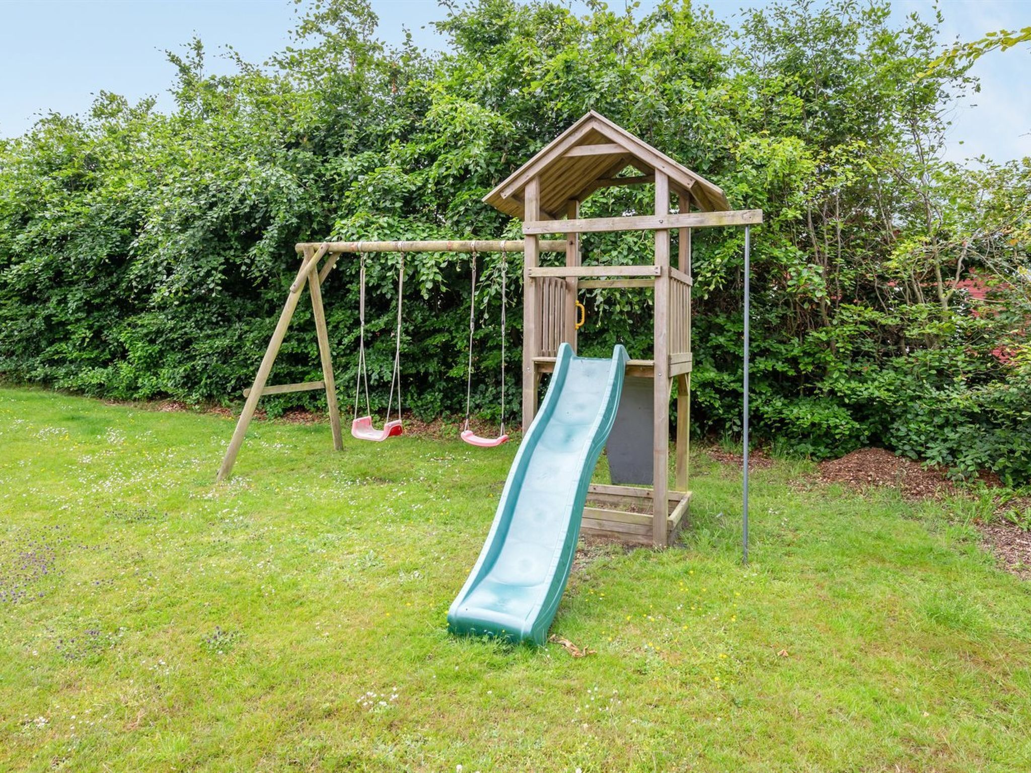 Een houten speelhuisje met een glijbaan en schommels in een grasveld.