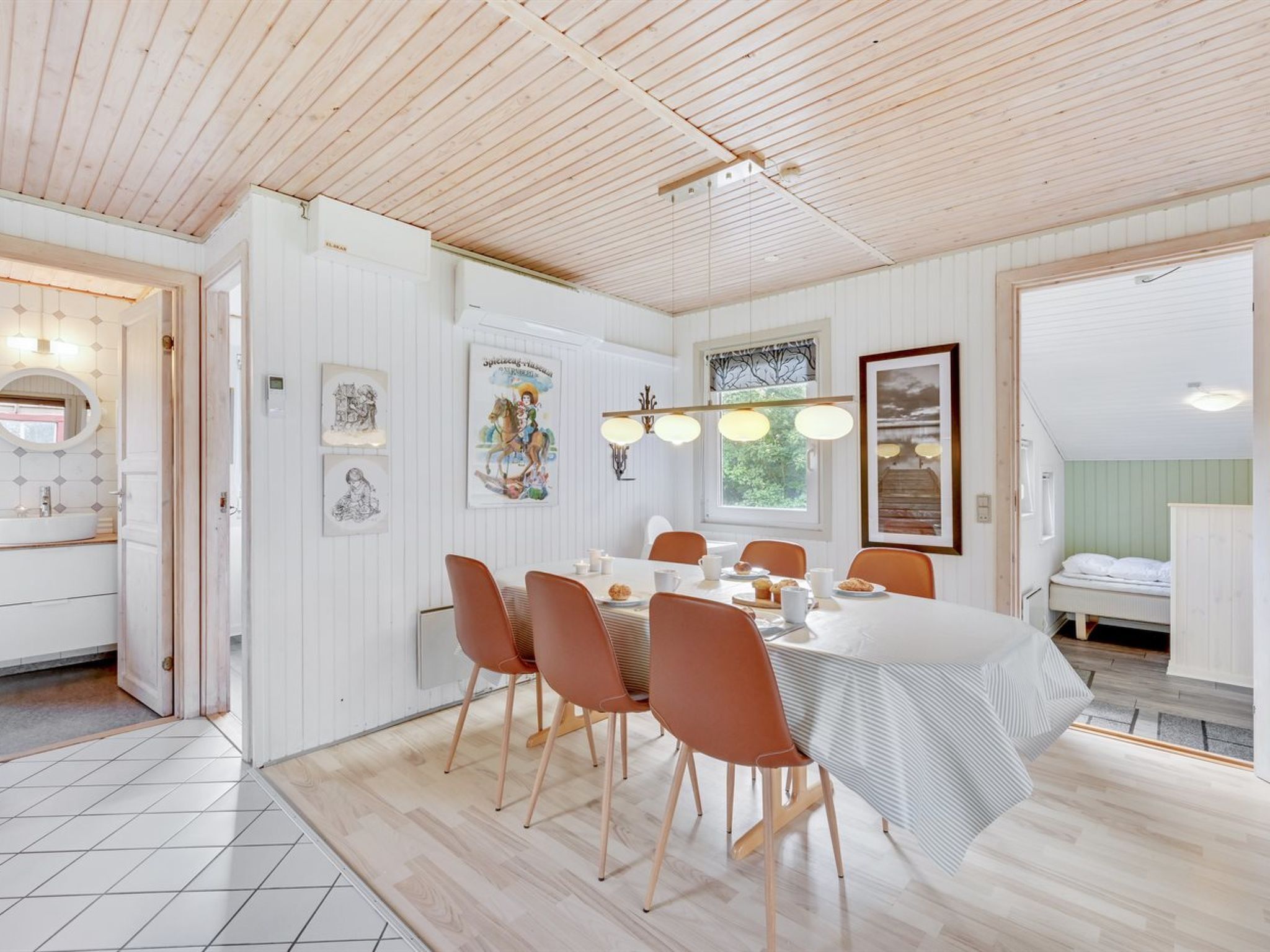 Un espace repas avec un plafond en bois, des murs blancs et un sol carrelé. On y trouve six chaises disposées autour d’une table recouverte d’une nappe blanche.