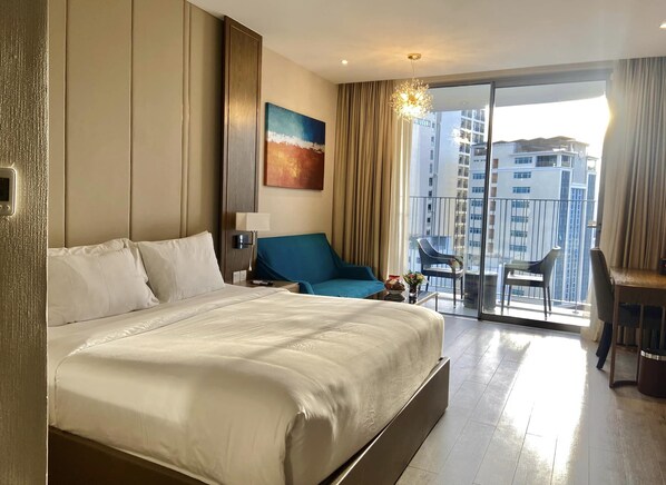 A hotel room with a large bed, a blue armchair, a small table with a vase, and a balcony with chairs.