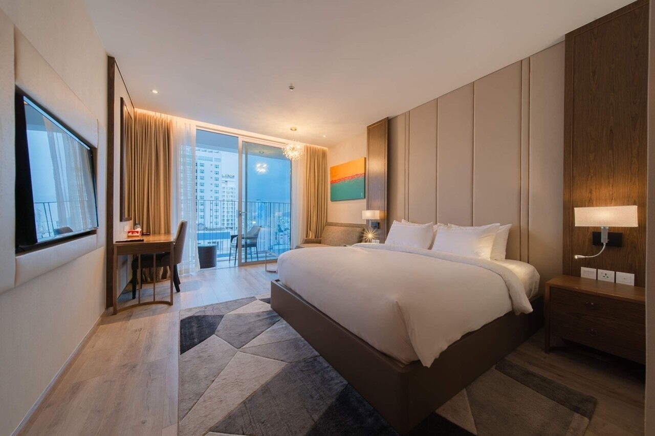 A hotel room with a large bed, a desk, a chair, a nightstand, and a view of the city through a large window.