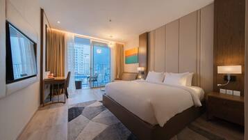 A hotel room with a large bed, a desk, a chair, a nightstand, and a view of the city through a large window.