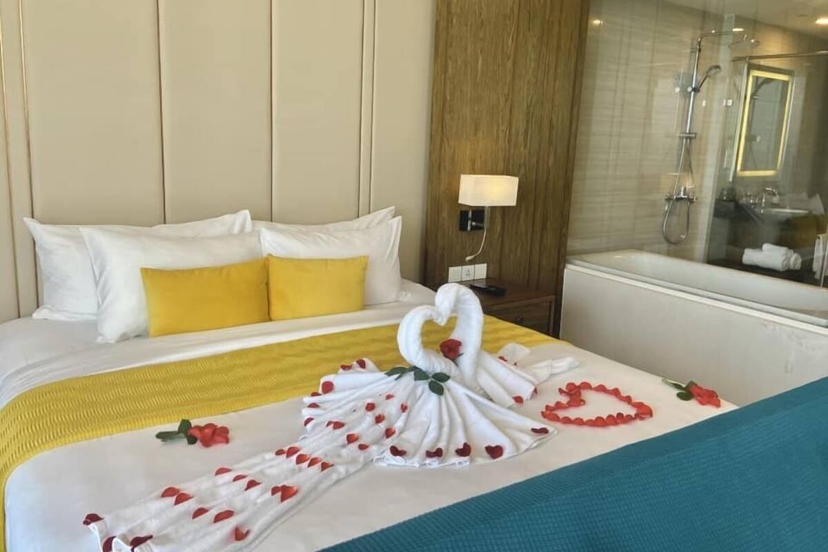 Ein Hotelzimmer mit einem ordentlich bezogenen Bett, einem hölzernen Kopfteil, einer Nachttischlampe sowie einem Badezimmer mit Dusche und Spiegel.