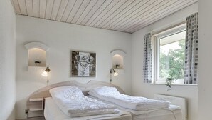 Uma cama de casal com cabeceira, luminárias de parede, uma obra de arte pendurada e uma janela com cortinas.
