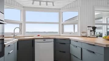 A modern kitchen with a dishwasher, a coffee maker, and a view of the sea through large windows.