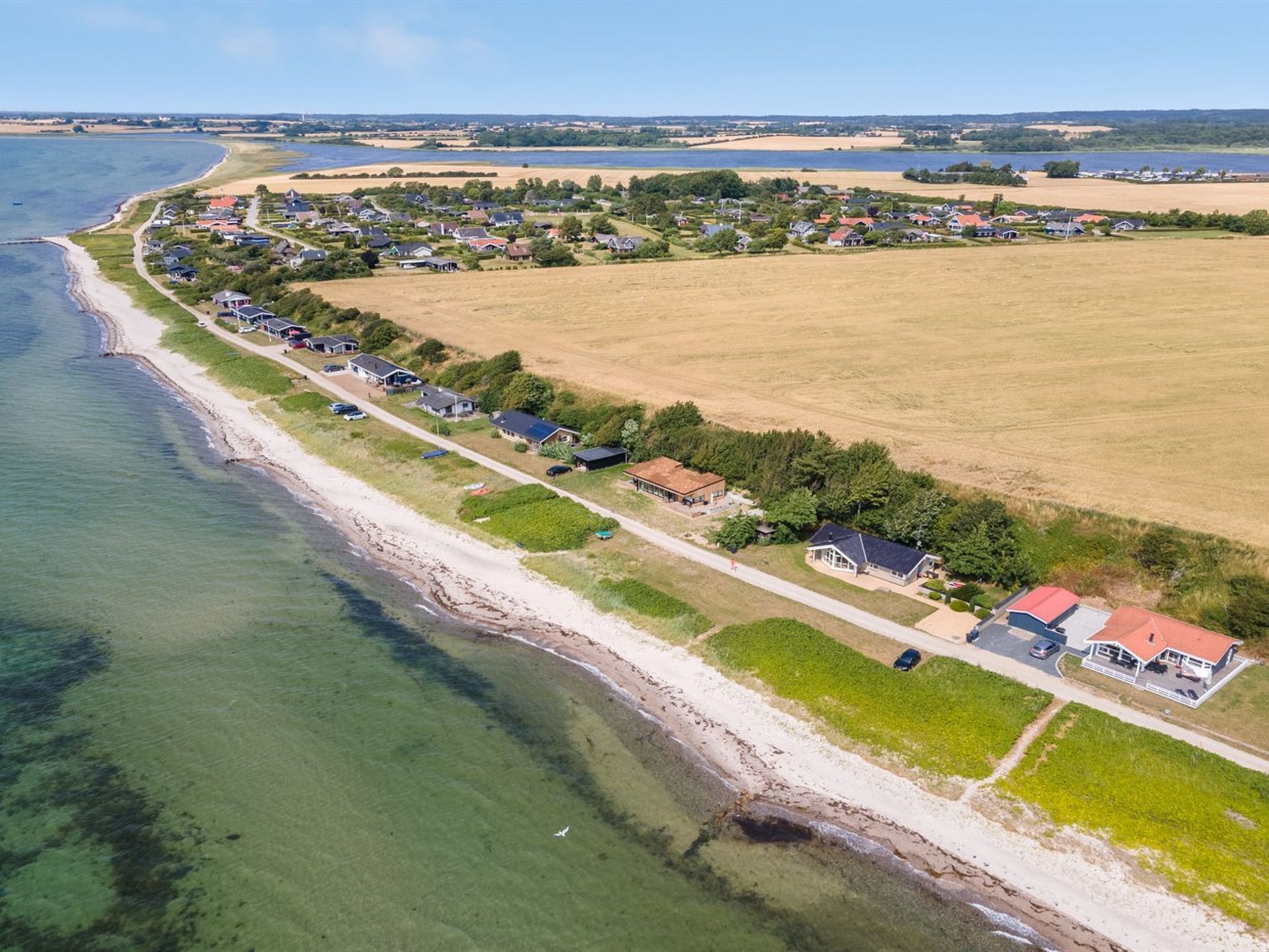 An aerial view of a coastal residential area with houses along the shoreline, a sandy beach, and open fields.