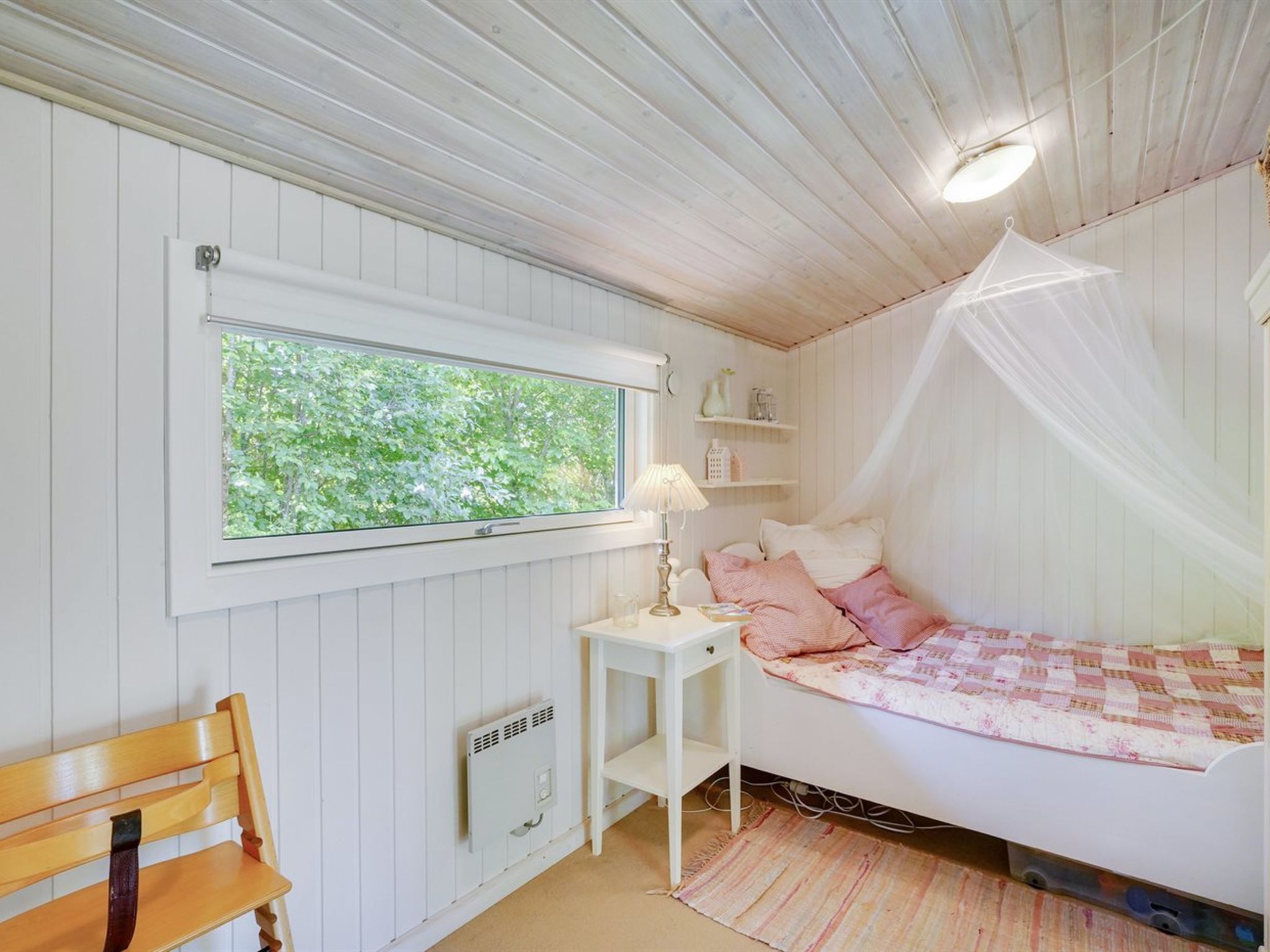 Makuuhuoneessa on sänky, yöpöytä, lamppu, penkki ja ikkuna, josta avautuu näkymä puihin.