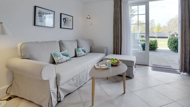 Een moderne woonkamer met een grijze bank, twee kussens, een bijzettafeltje met een schaal fruit en een ingelijst schilderij aan de muur.