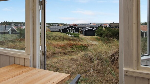 Vue depuis l’intérieur d’une pièce : on y aperçoit une table en bois et une fenêtre donnant sur un espace verdoyant où se trouvent plusieurs maisons.