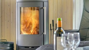 A modern wood-burning stove with a glass door, a bottle of wine, and two glasses on a glass table in a cozy room with wooden walls and a leather couch. - "Wegher" - 650m from the sea by Interhome (Væggerløse)