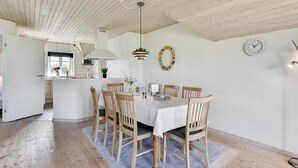 Et spiseområde med et træbord, seks stole, en lysekrone, et ur og et køkken i baggrunden.