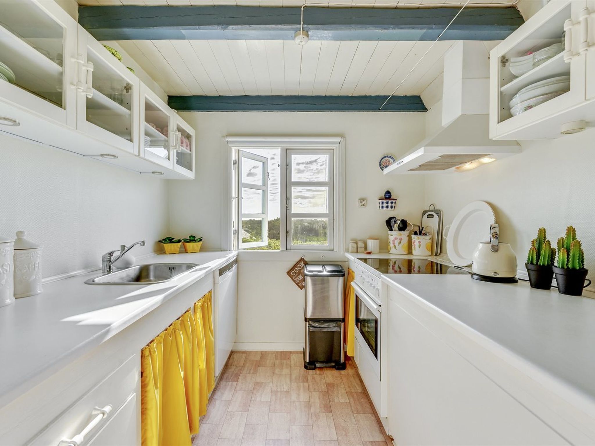 A compact kitchen with white cabinets, a stainless steel sink, a window, and a trash bin.