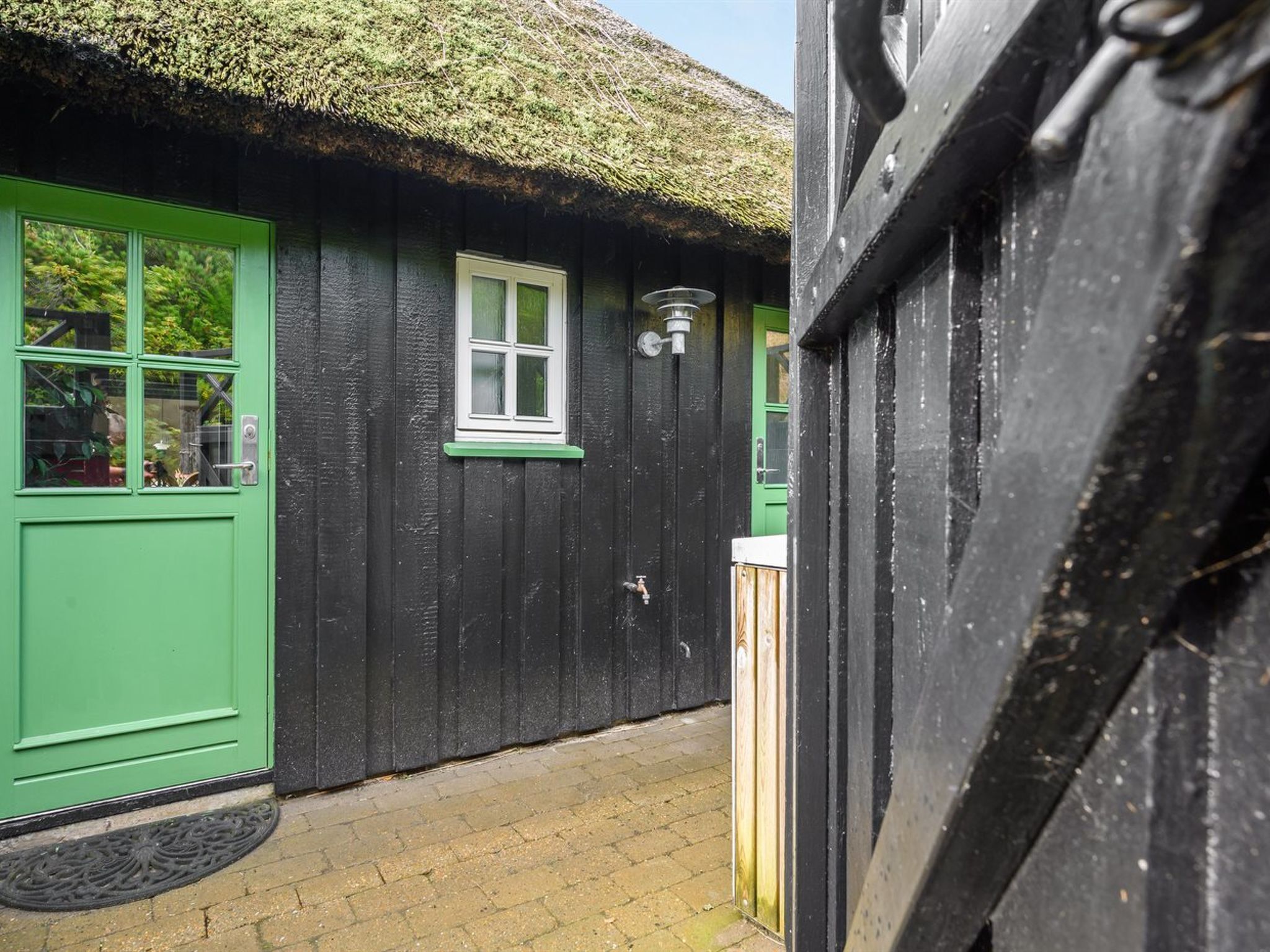 Un bâtiment en bois noir, avec une porte et des fenêtres vertes, un toit de chaume et une petite fenêtre dotée d’un luminaire.