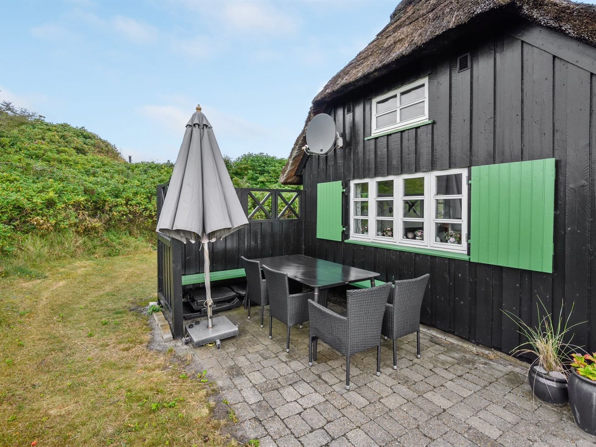 A wooden house with a thatched roof, a table and chairs outside, and a satellite dish on the wall.