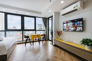A modern hotel room with large windows, a bed, a dining table with chairs, a TV mounted on the wall, and a small plant on the side table.