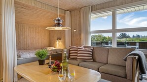 Una sala de estar acogedora, con techo de madera, un sofá con cojines a rayas, una mesa de centro con una botella y vasos, y vista a zonas verdes al exterior.