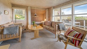Una habitación acogedora con paredes y techo de madera, un sofá, una silla, una mesa y ventanas amplias que ofrecen vistas al exterior.