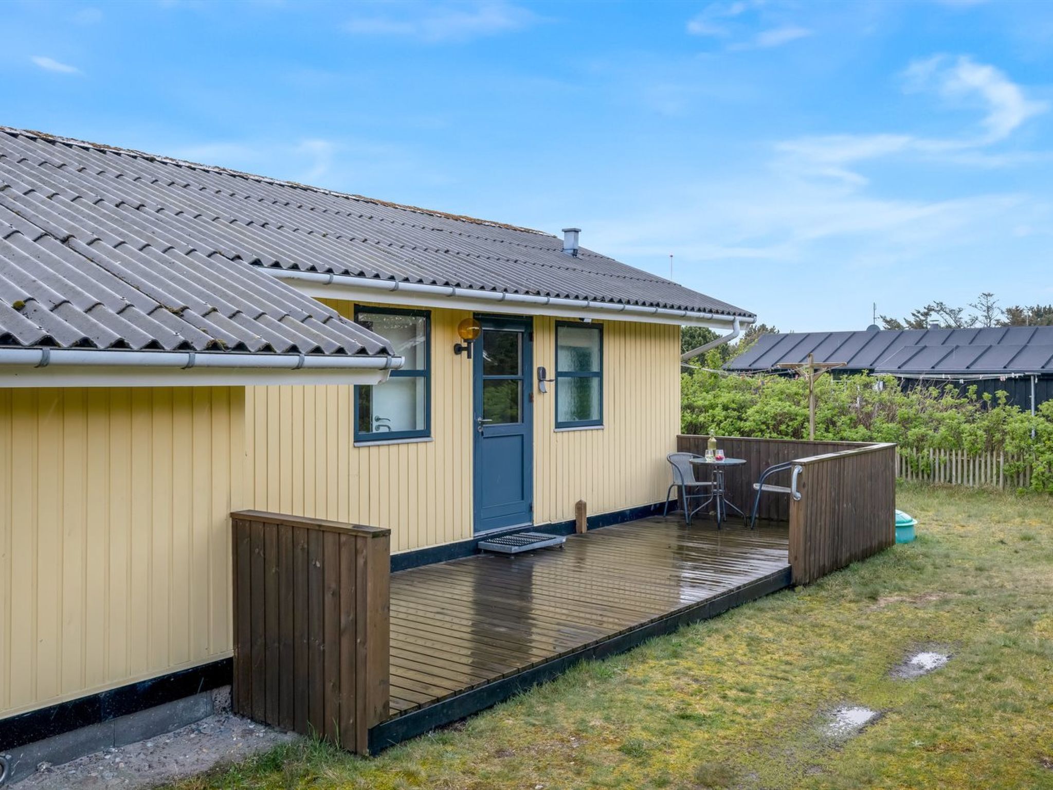 Een geel gebouw met een grijs dak, een houten terras en een klein patio met een tafel en stoelen.