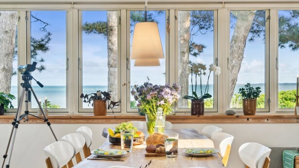 A dining area with a table set for four, large windows with a view of the sea, and potted plants on the windowsill.
