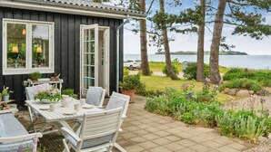 A cozy outdoor seating area with a table and chairs, a small garden with various plants and flowers, and a view of the sea and trees in the background.