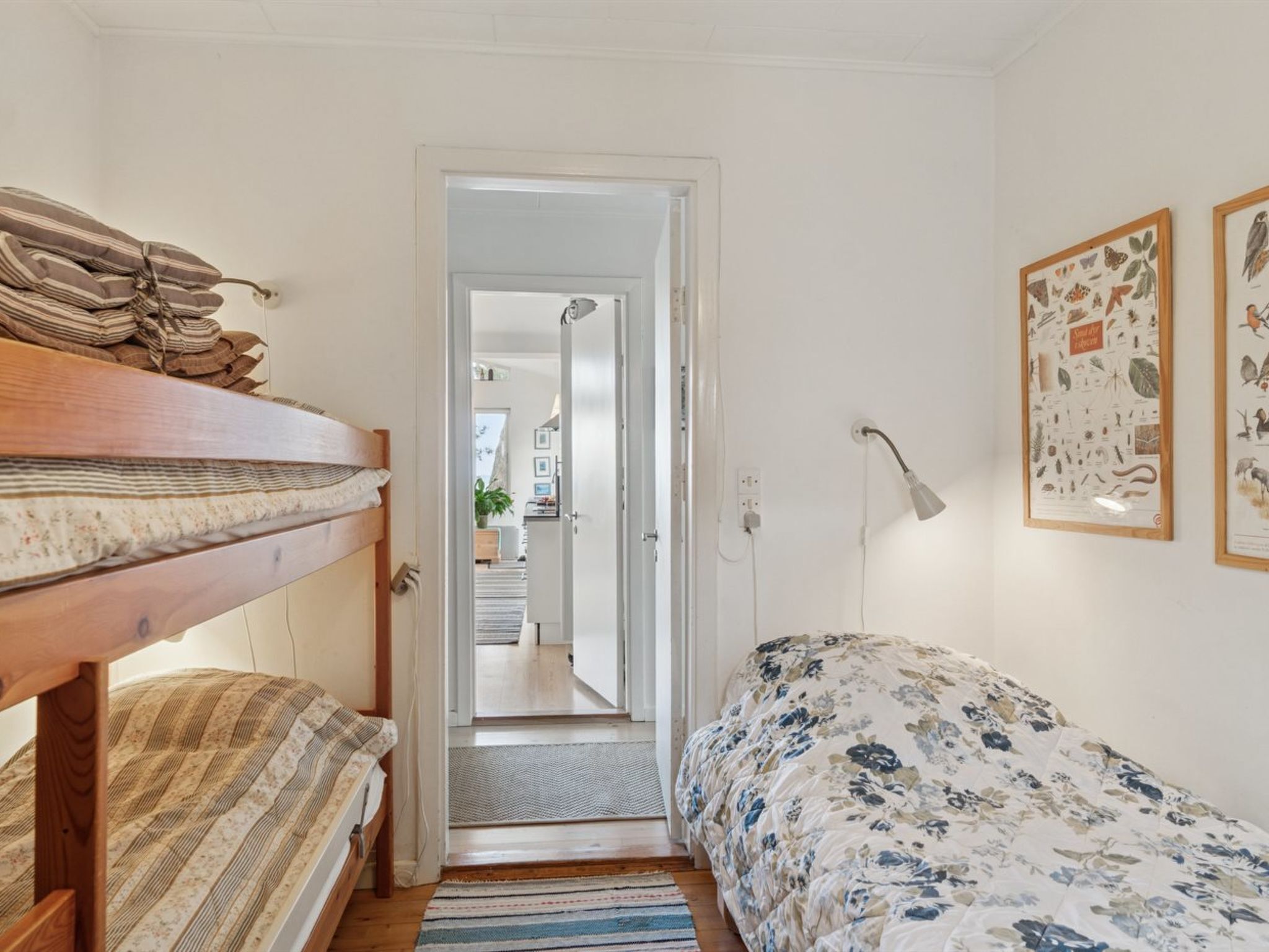 Un dormitorio con una cama litera, una cama individual, una puerta que da a un pasillo y dos láminas botánicas enmarcadas en la pared.