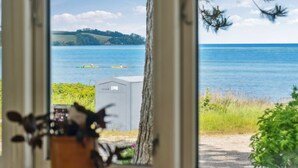 A view through a window of a coastal scene with kayaks on the water, a small shed, and greenery.